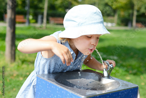 Cute baby girl drinking from water drinking fountain in summer