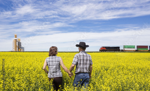 horizontal image of a husband and wife checking canola field while a train rumbles by in the background next to a grain elevator.
