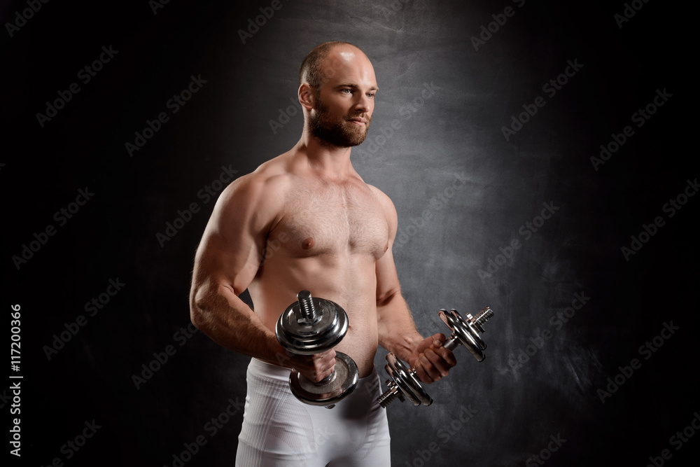 Young powerful sportsman training with dumbbells over black background.