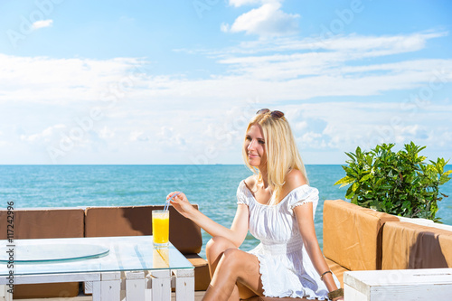 Young smiling and happy woman drinking juice in beach cafe with sky and sea background.