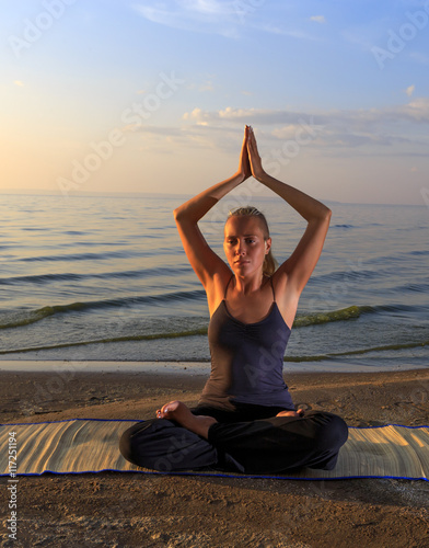 Beautiful young woman practising yoga on mat outdoors at river bank on sand at sunset