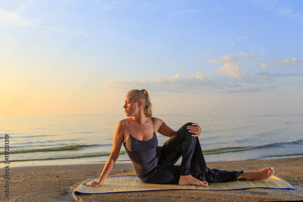 Beautiful young woman practising yoga on mat outdoors at river bank on sand at sunset