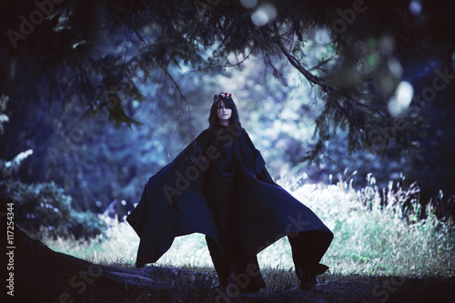 Young black-haired girl in long cloak with a hood in dark forest. Effect of toning photo