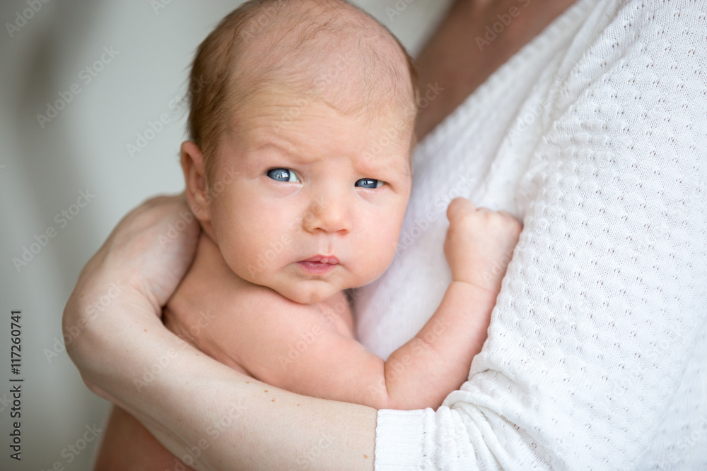 Portrait of cute newborn baby looking at camera with funny serious expression while sitting on mothers arms. Young mom holding healthy new born child. Love, childcare, happy family concepts Portrait of cute newborn baby looking at camera with funny serious expression while sitting on mothers arms. Young mom holding healthy new born child. Love, childcare, happy family concepts