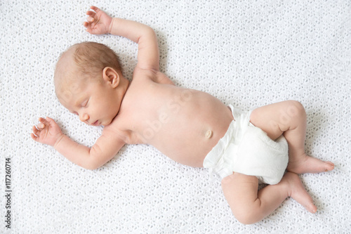 Portrait of young funny newborn babe napping on white knitted blanket with his arms up. Cute new born child sleeping. Healthy kid in diaper lying on bed with closed eyes. Full length. View from above