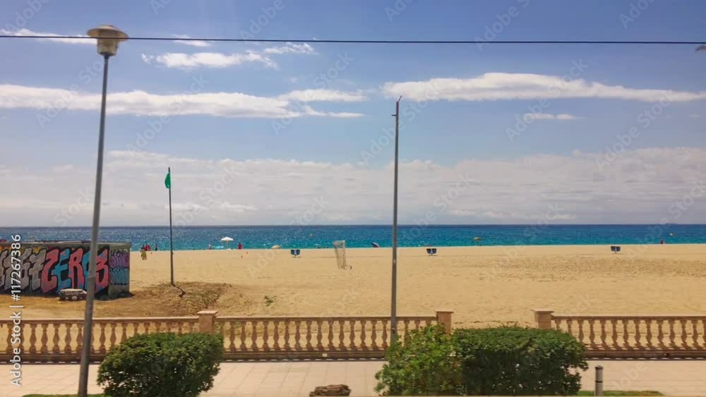 Costa Brava Spain Catalonya seashore, a view from train on long mediterranean sea coastline with palms, people swimming and taking sun baths, waves crashing over sand beach, train stops at station