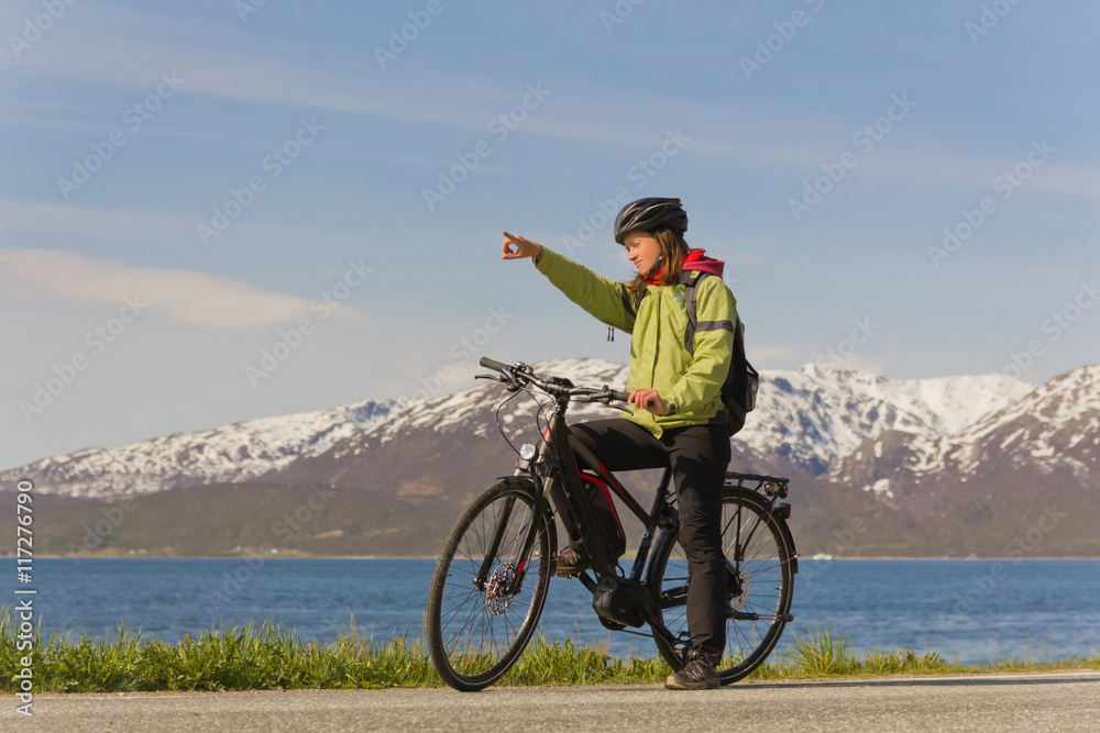 Obraz premium Frau auf E-Bike/ Fahrrad zeigt den Weg. Sie steht mit ihrem Pedelec vor einem Bergpanorama und Wasser. Woman on electric bike / bicycle showing the way