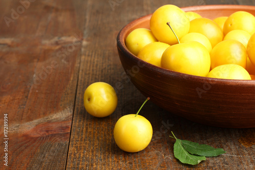 Yellow plums in a bowl on brown wooden background.