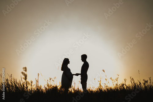 Silhouette portrait of beautiful young anonymous pregnant family in expectation of baby. Man and woman standing face to face over sunset sky background in evening in scenic nature landscape background
