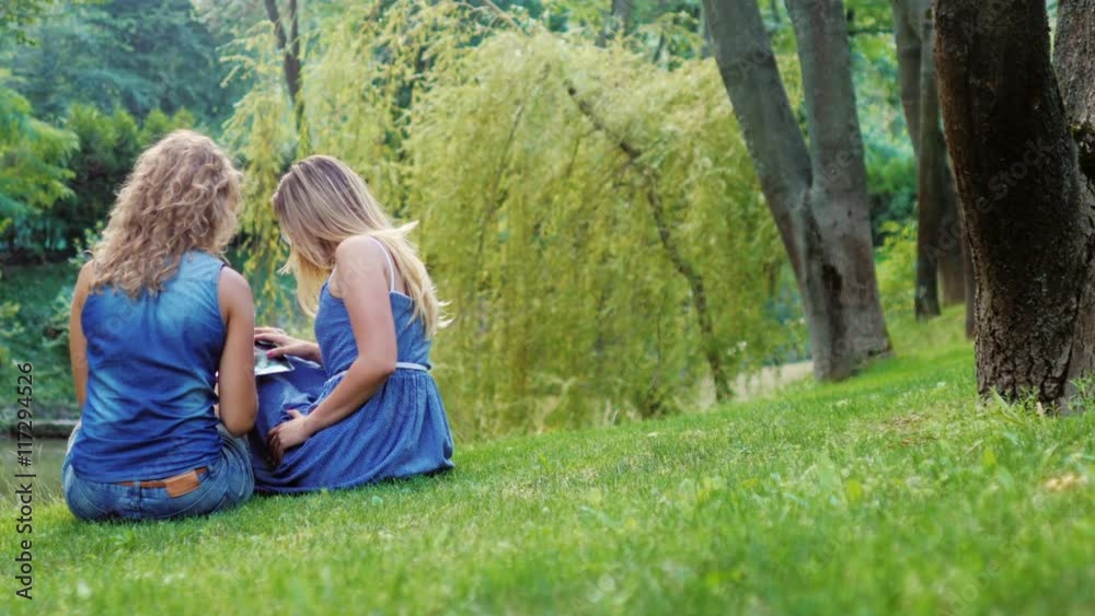 Two friends - the young woman relaxing on the green grass in the park. enjoy tablet. Back view Two friends - the young woman relaxing on the green grass in the park. enjoy tablet. Back view
