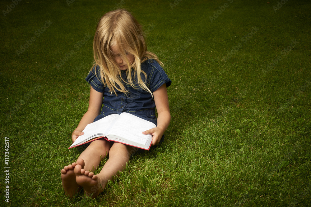 Fototapeta premium Cute Little Blond Girl Reading Book Outside on Grass