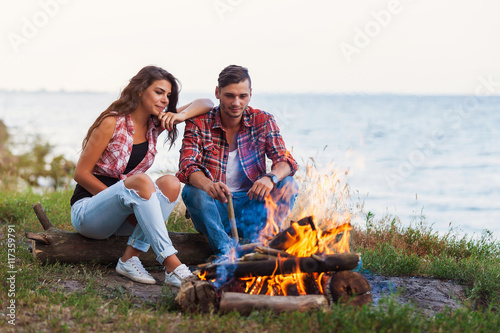 friendship, happiness, summer vacation, holidays and people concept - close up of couple sitting near fire on beach