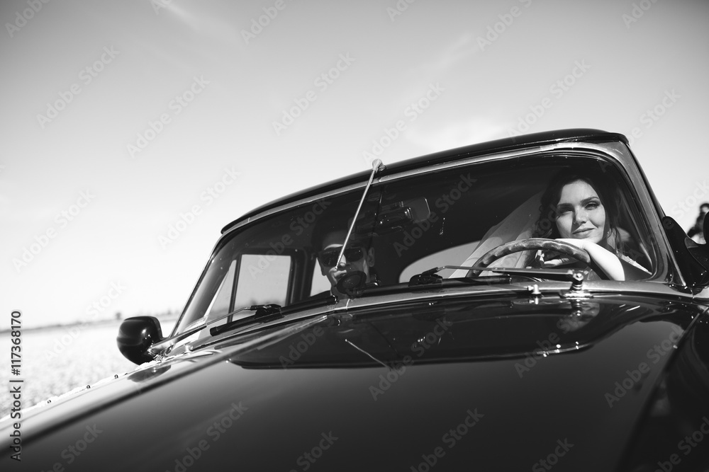 Smiling newlyweds sit in an old car in a sunny day