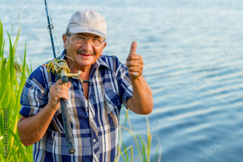 a glad fisherman with a fishing rod and his thumb up by the river on a nice sunny day. Everything is OK!