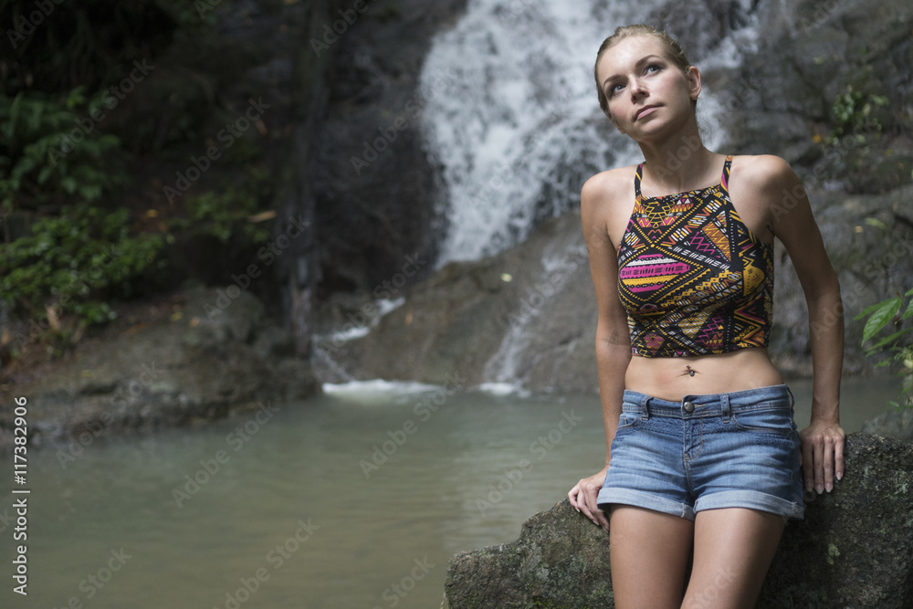 Pretty blonde woman sittting on a rock and looking up near waterfall. Female tourist enjoying by a water fall in forest