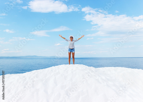 Young woman standing on white beach with her arms outstretched over horizon. She is looking towards the sun.