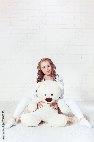 Tender blonde girl wearing in wight clothes hugging teddy bear on brick wall background. Girl with brown eyes looking at the camera image released.