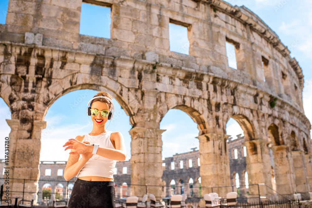 Young sports woman with headphones and smartwatch resting after the training near the ancient coliseum in Pula city