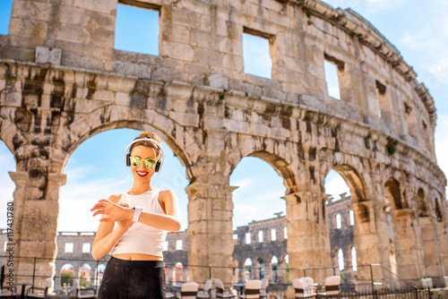 Young sports woman with headphones and smartwatch resting after the training near the ancient coliseum in Pula city