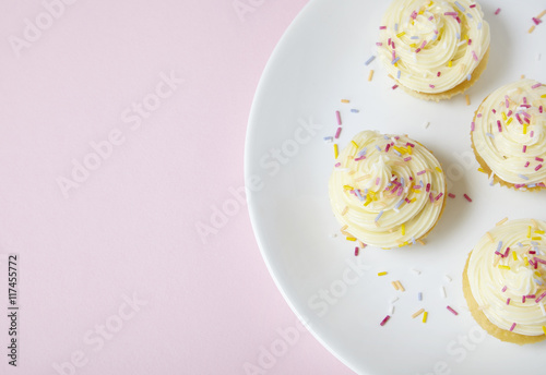 A plate of birthday cupcakes with vanilla frosting and sprinkles on a pastel pink background