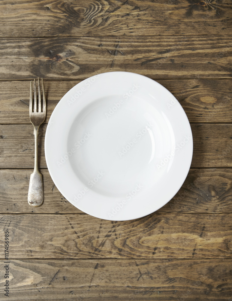 An empty white dinner plate and fork on a rustic wooden background