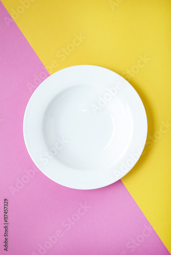 An empty white dinner plate on a two toned pink and yellow background