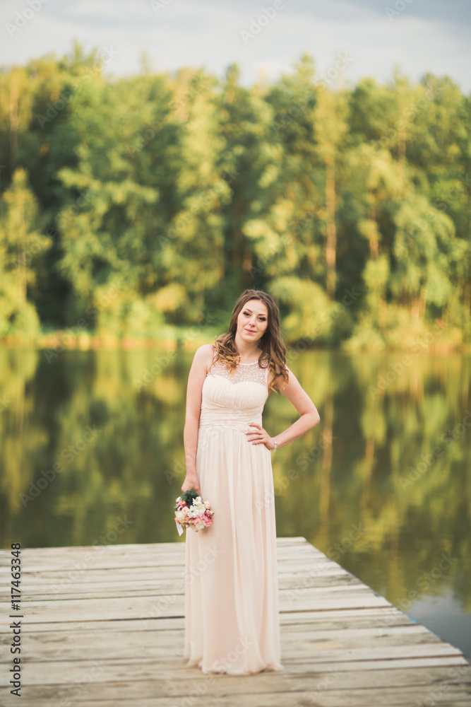 Gorgeous bride in elegant dress holding bouquet posing near forest and lake Gorgeous bride in elegant dress holding bouquet posing near forest and lake