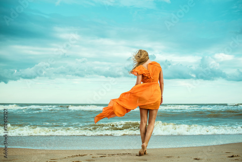 A young slender woman in orange dress is walking barefoot towards the storming sea. The train of her dress is waving. Cool wind in her hair. Outdoor shot. Copy-space.