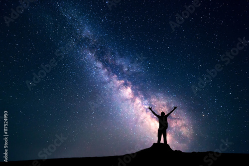 Landscape with colorful Milky Way. Night sky with stars and silhouette of a happy man with backpack and raised up arms on the stone. Space background