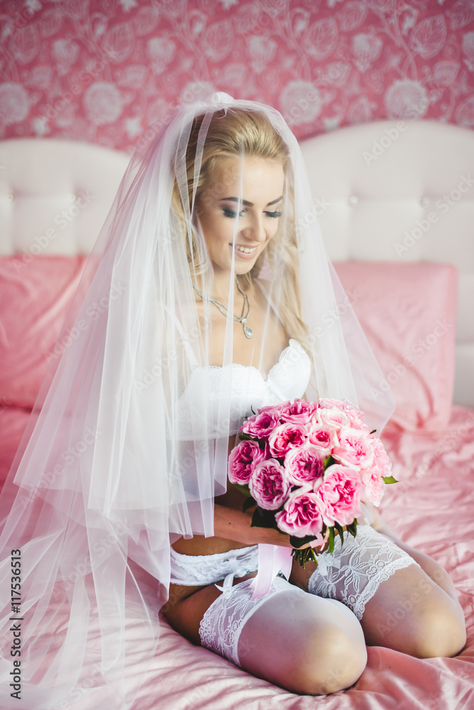 Beautiful sexy young bride underwear sitting on a bed with a bouquet of flowers in hands and with a veil on her head. Morning the bride. Beautiful sexy young bride underwear sitting on a bed with a bouquet of flowers in hands and with a veil on her head. Morning the bride.