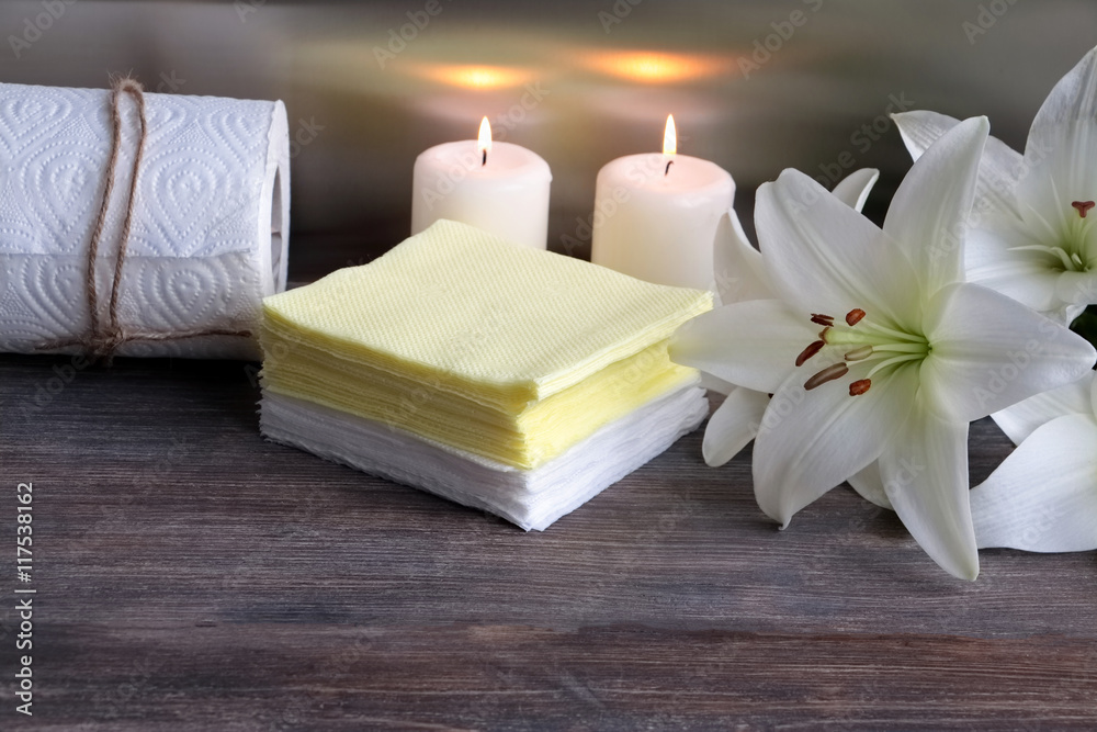 White and yellow paper napkins,paper towel on wooden table against steel background with lily flowers and burning candles. Concept of spa and body care