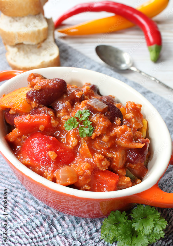 chili con carne mit chillies, frischem Gemüse, Petersilie und Gewürzen in einer kleinen orangenen Schüssel mit Löffel und Brot auf einem grauen Tischtuch
