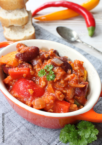 chili con carne mit chillies, frischem Gemüse, Petersilie und Gewürzen in einer kleinen orangenen Schüssel mit Löffel und Brot auf einem grauen Tischtuch