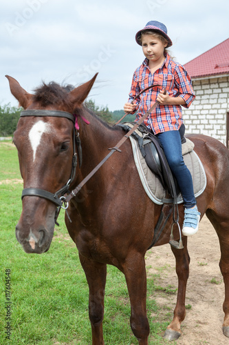 Young girl in a plaid shirt and hat sitting on horseback