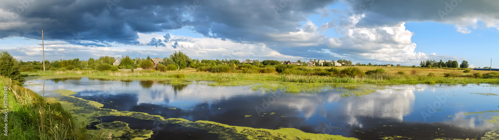 Village settlement. Rural landscape panoramic. Reflection of large clouds in the water. Panorama of idyllic lakes and clouds to the horizon. Country living in the houses.