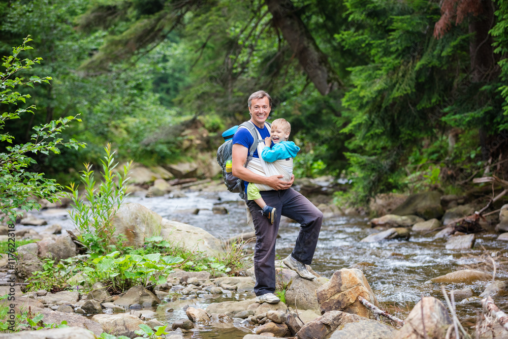 Caucasian man carrying his son in wrap sling while hiking near mountainous river 
