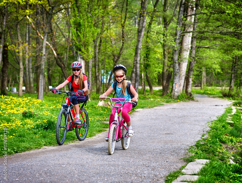 Fototapeta premium Girls wearing bicycle helmet and glasses with rucksack rides bicycle on asphalt track. Children ride on green grass and flowers in park.