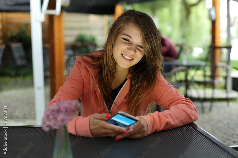 Uśmiechnięta dziewczyna z telefonem przy stoliku w restauracji. - obrazy, fototapety, plakaty 