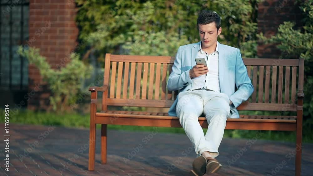 Casual businessman texting on phone on park bench on a sunny day Casual businessman texting on phone on park bench on a sunny day