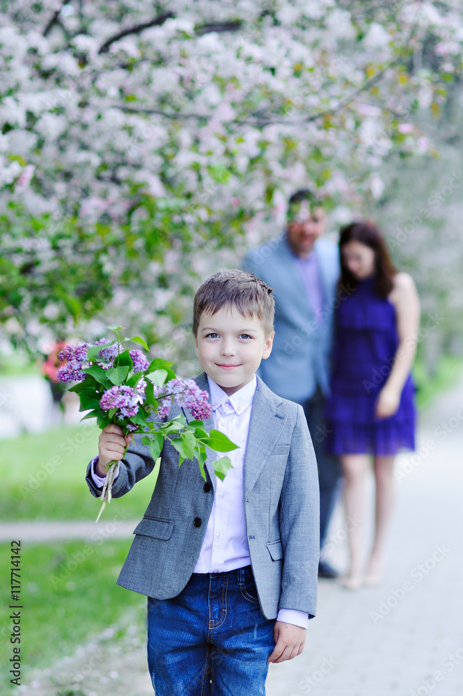 Fototapeta premium The boy goes with the bouquet of lilacs blooming along the alley, in the background are embracing his parents, the look in the picture.
