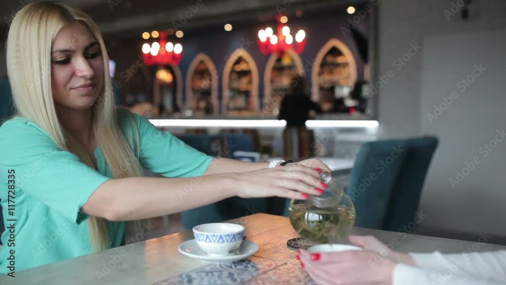 The blonde pours a cup of tea from a teapot.