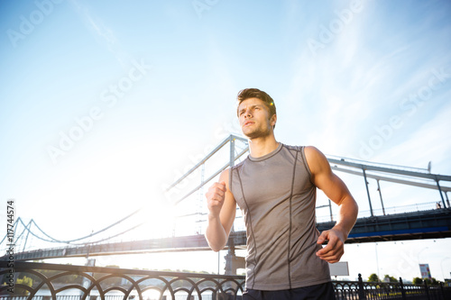 Handsome sports man running along modern bridge at sunset light