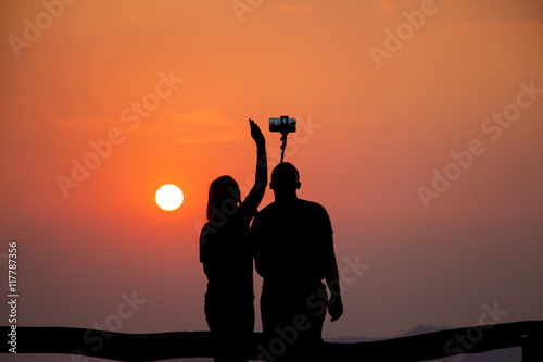 A tourist couple is taking selfies against the setting sun as seen from their dark silhouettes