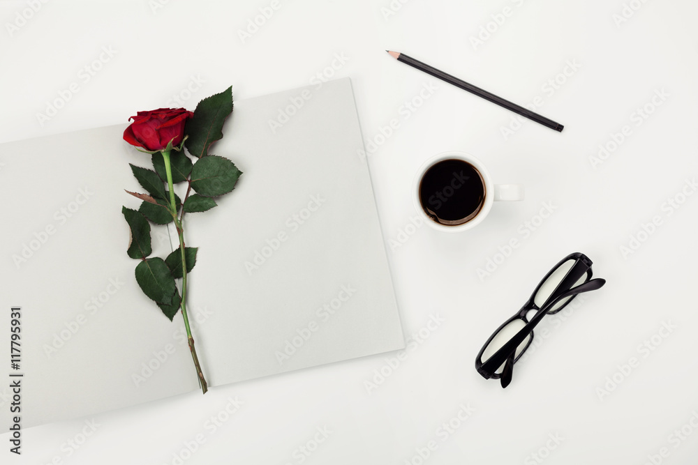 Womens working space with cup of coffee, pencil, empty notebook, glasses and rose flower on white table overhead view. Flat lay styling. Womens working space with cup of coffee, pencil, empty notebook, glasses and rose flower on white table overhead view. Flat lay styling.