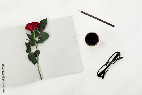 Womens working space with cup of coffee, pencil, empty notebook, glasses and rose flower on white table overhead view. Flat lay styling.
