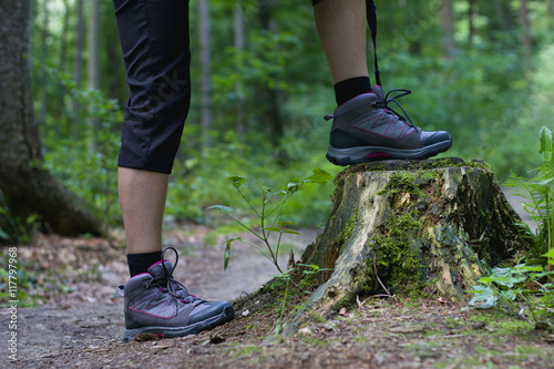 A young woman standing on a tree stump in the middle of forest trail, hiking boots in focus