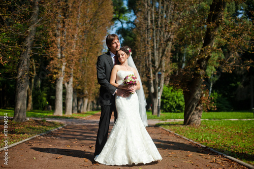 Groom lies his chin over bride's head hugging her in the park photo