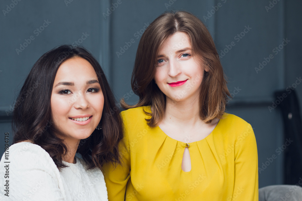 Caucasian and Asian young women are sitting on the couch in beautiful dresses Caucasian and Asian young women are sitting on the couch in beautiful dresses