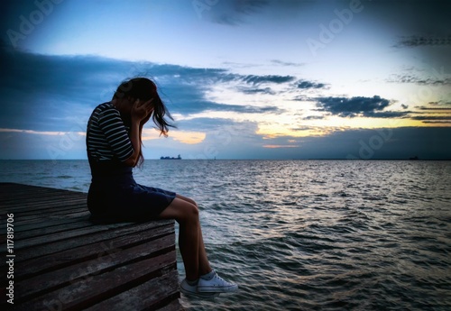 Beautiful woman in frustrated depression sitting on wooden bridge, near the beach on sunset. Concept of unemployed, sadness, depressed and human problems in dark tone.