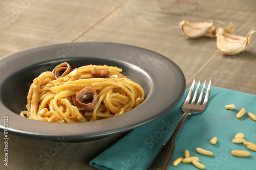 A grey dish of spaghetti with breadcrumbs, anchovies, garlic and pine nuts. A fork and some pine nuts on a blue cotton napkin. Background: garlic on a wooden board.